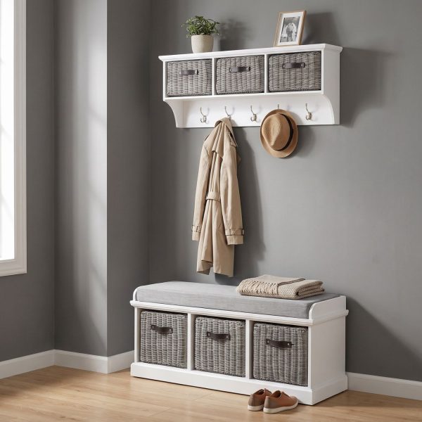 A white wall-mounted shelf unit displaying a potted plant and a small framed photo on top, with three grey faux rattan baskets featuring dark handles in its cubbies. Five silver double-hooks below hold a beige trench coat and a straw hat with a black band. Directly below sits a matching white wooden storage bench topped with a grey cushion and a folded throw blanket, containing three larger matching grey faux rattan baskets in its base. The furniture is styled against a smooth grey wall with light wood flooring and a pair of brown shoes resting beside the bench.