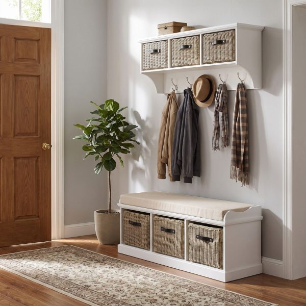 A white wall-mounted shelf unit displaying small decorative boxes on top, with three natural woven baskets featuring dark handles. Five silver double-hooks below hold a tan jacket, a dark jacket, a straw hat, and two patterned scarves. Directly below sits a matching white wooden storage bench topped with a beige cushion, containing three larger matching natural woven baskets in its base. The furniture is styled in a bright entryway next to a wooden door and a potted fiddle leaf fig plant, with wood flooring and a patterned runner rug.
