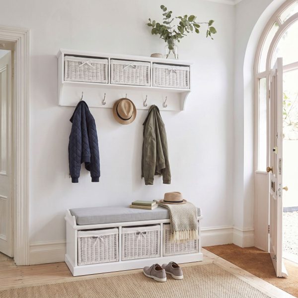 A white wall-mounted shelf unit displaying a glass vase with leafy branches on top, with three white woven baskets featuring small white bow details in its open compartments. Five silver double-hooks below hold a navy quilted jacket, a straw hat, and an olive green jacket. Directly below sits a matching white wooden storage bench topped with a grey cushion, a couple of books, another straw hat, and a draped fringed blanket. The bench's base contains three larger matching white woven baskets. The furniture is styled in a bright entryway next to an open arched white door, with light wood flooring, a woven rug, and a pair of grey casual shoes resting beside the bench.