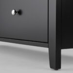 Extreme close up of the legs attatched to the bottom of a black chest of drawers, photographed against a white background.